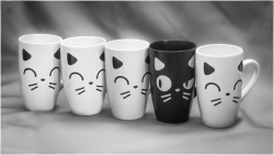 Five ceramic mugs with stylised cat faces sit side by side; four are white with matching closed‑eye expressions, while the central black mug features one open eye, creating a playful contrast
