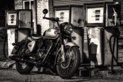 A vintage Royal Enfield motorcycle stands in front of three weathered, rusting petrol pumps, captured in high‑contrast black and white. (Monochrome image)