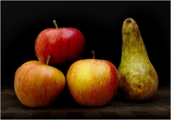 Still life of three red apples with yellow tones arranged in a triangle beside an upright speckled pear, all set on a dark wooden surface with a deep black backdrop