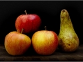 Still life of three red apples with yellow tones arranged in a triangle beside an upright speckled pear, all set on a dark wooden surface with a deep black backdrop