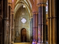 Interior view of a Gothic church corridor with pointed arches, ribbed ceilings, ornate columns, and coloured light from stained‑glass windows illuminating the stone floor.