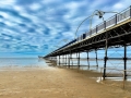 Long pier stretching over a sandy beach at low tide, with metal supports and dramatic cloud‑filled sky.
