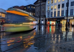 A tram moves through a city street at dusk, captured with long‑exposure light trails reflecting on wet pavement beside illuminated shopfronts. (Colour image)