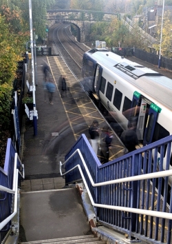 Passengers move around a Dronfield train station platform as a white‑and‑blue train waits beside autumn trees and a stone bridge, viewed from the top of a staircase. (Colour image)