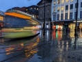 A tram moves through a city street at dusk, captured with long‑exposure light trails reflecting on wet pavement beside illuminated shopfronts. (Colour image)