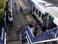 Passengers move around a Dronfield train station platform as a white‑and‑blue train waits beside autumn trees and a stone bridge, viewed from the top of a staircase. (Colour image)