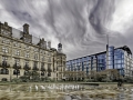 Urban square with a historic stone building, modern glass tower, and circular fountain under dramatic clouded skies.