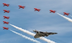 A large grey delta‑wing aircraft flies at the centre of a tight formation, surrounded by eight smaller red jets that trail white smoke across a clear blue sky