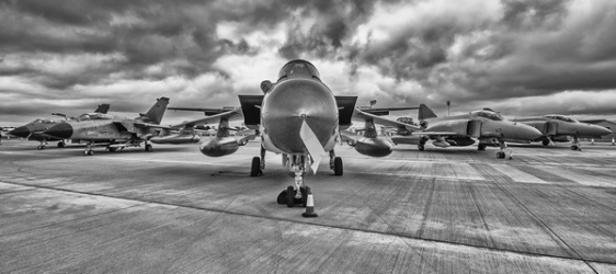 Black‑and‑white line up of fighter jets on a runway beneath a dramatic cloudy sky.