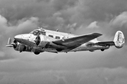 A polished metal twin‑engine aircraft flies against a cloudy sky, its mid‑20th‑century design highlighted by spinning propellers and reflective surfaces. (Monochrome image)