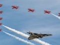 A large grey delta‑wing aircraft flies at the centre of a tight formation, surrounded by eight smaller red jets that trail white smoke across a clear blue sky