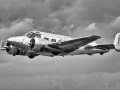 A polished metal twin‑engine aircraft flies against a cloudy sky, its mid‑20th‑century design highlighted by spinning propellers and reflective surfaces. (Monochrome image)
